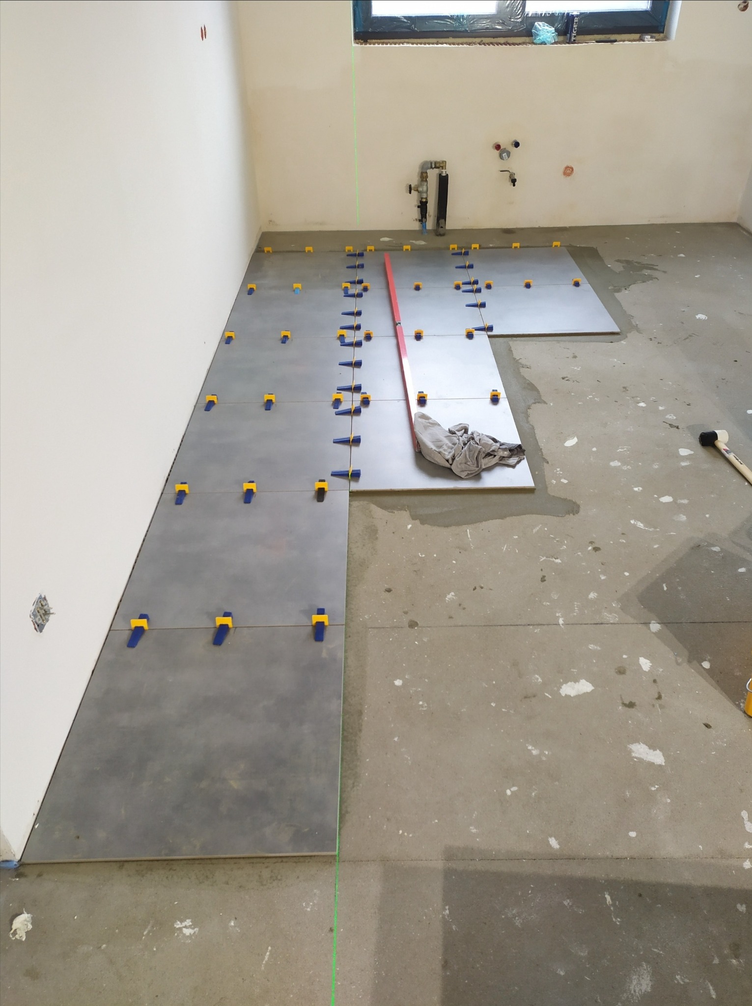 Układanie szarych płytek podłogowych w surowym wnętrzu z użyciem poziomicy laserowej i klipsów poziomujących, widoczny gumowy młotek i szmatka.
