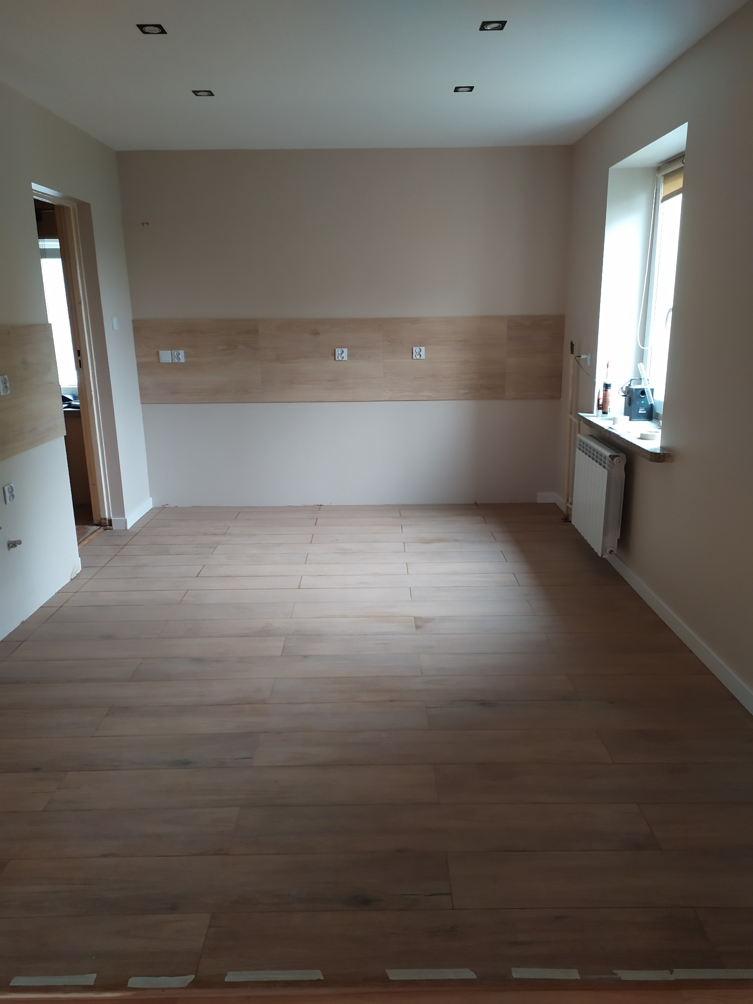 Puste pomieszczenie po remoncie z drewnopodobną podłogą, fragmentem ściany wyłożonym płytkami i grzejnikiem pod oknem, gotowe do umeblowania.