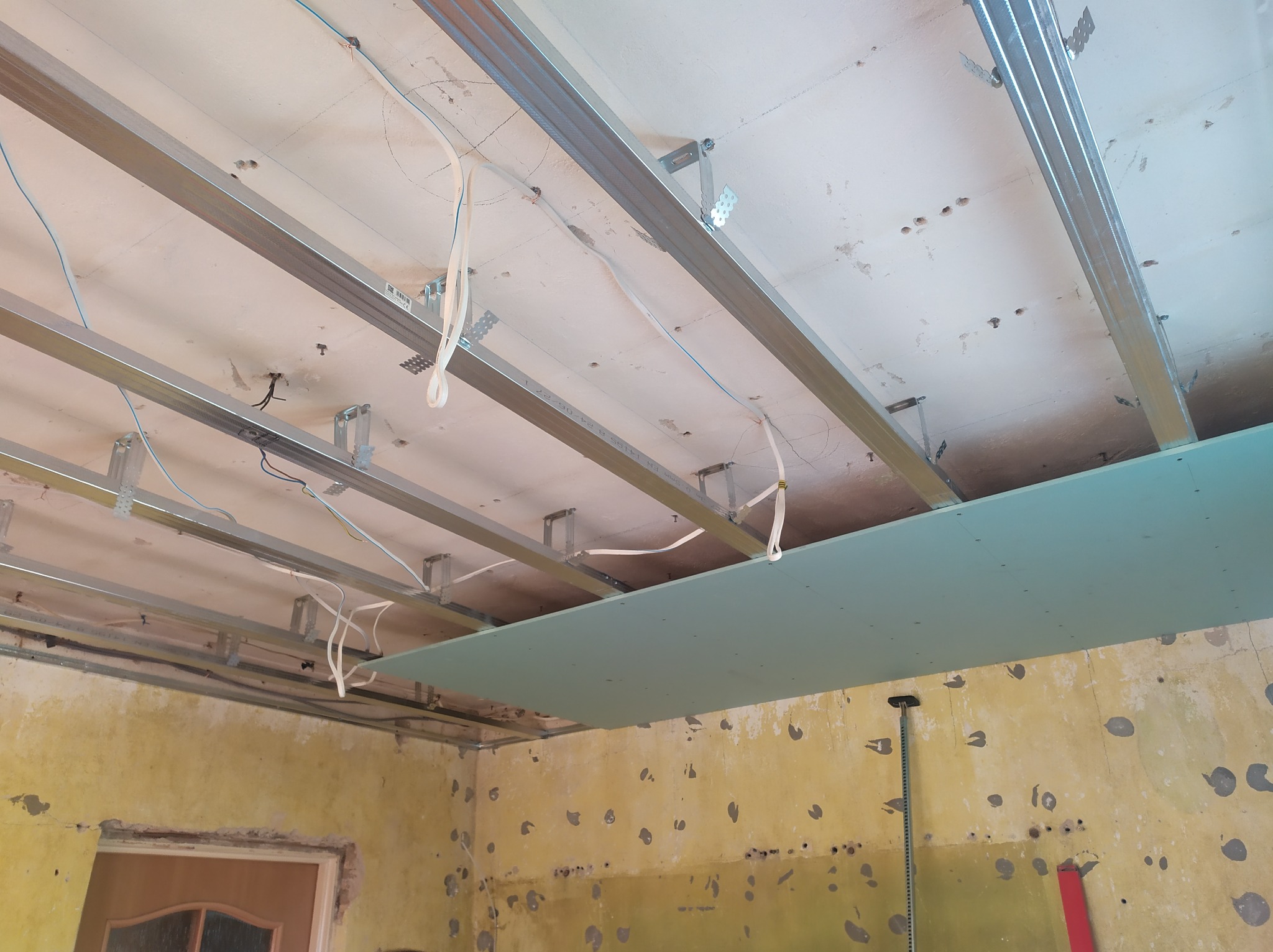 Montaż metalowej konstrukcji sufitu podwieszanego z płyt gipsowo-kartonowych w pomieszczeniu z widocznymi śladami wcześniejszych prac, żółtą ścianą i wystającymi kablami.