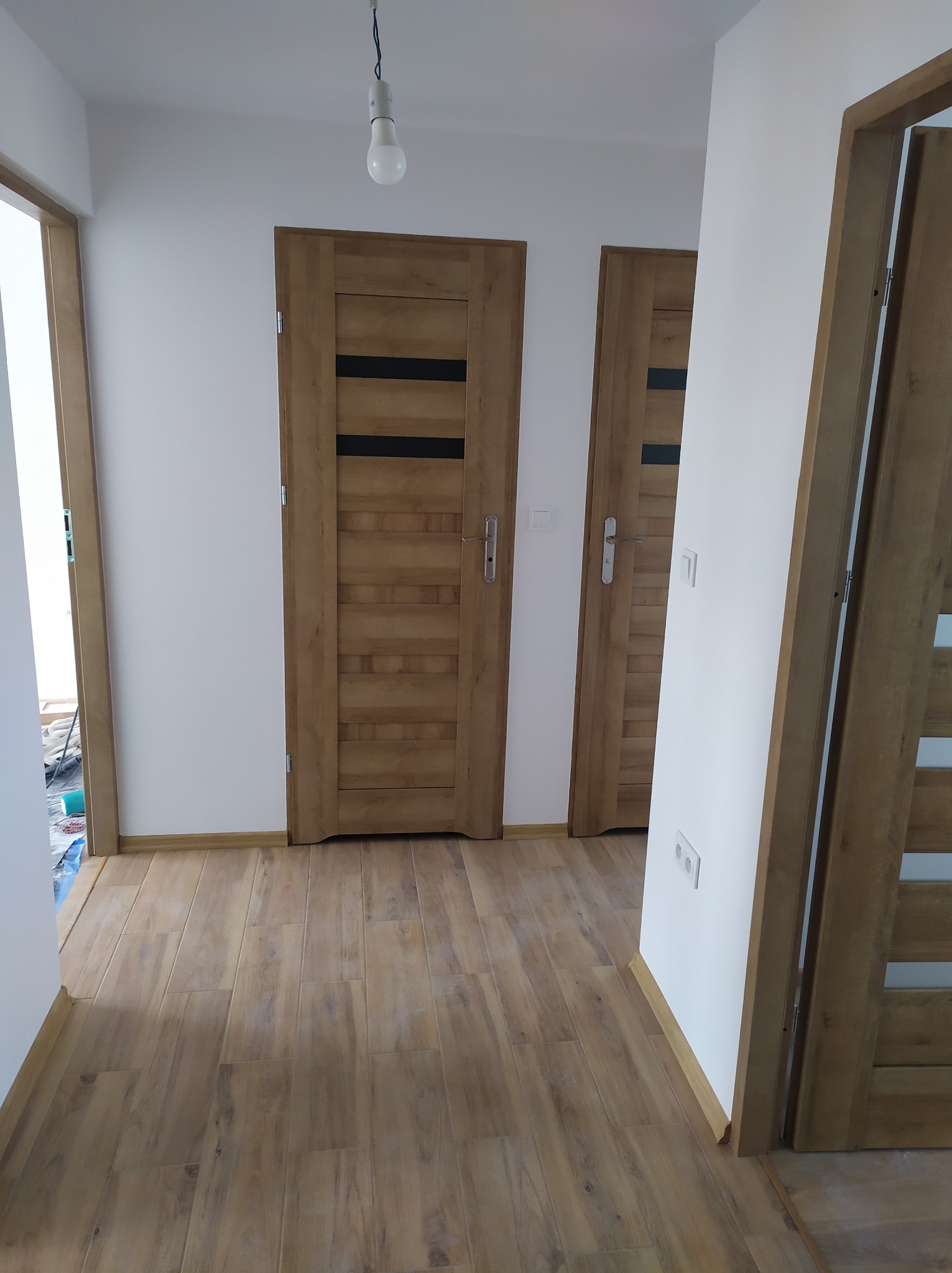 Korytarz po remoncie z drewnopodobną podłogą, białymi ścianami i trzema drewnianymi drzwiami z czarnymi wstawkami, oświetlony wiszącą żarówką.