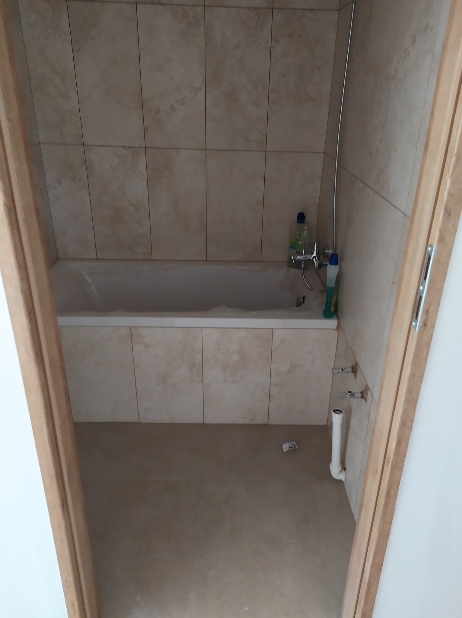 Wykończona łazienka z beżowymi płytkami, białą wanną, baterią i widocznymi rurami odpływowymi, sfotografowana zza drewnianej futryny.