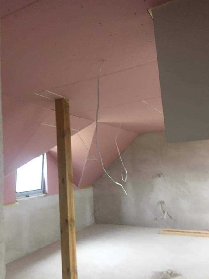 Poddasze w trakcie ocieplania, widoczne różowe płyty izolacyjne na skośnym suficie, drewniany słup podtrzymujący konstrukcję, wystające kable i okno w ścianie szczytowej.
