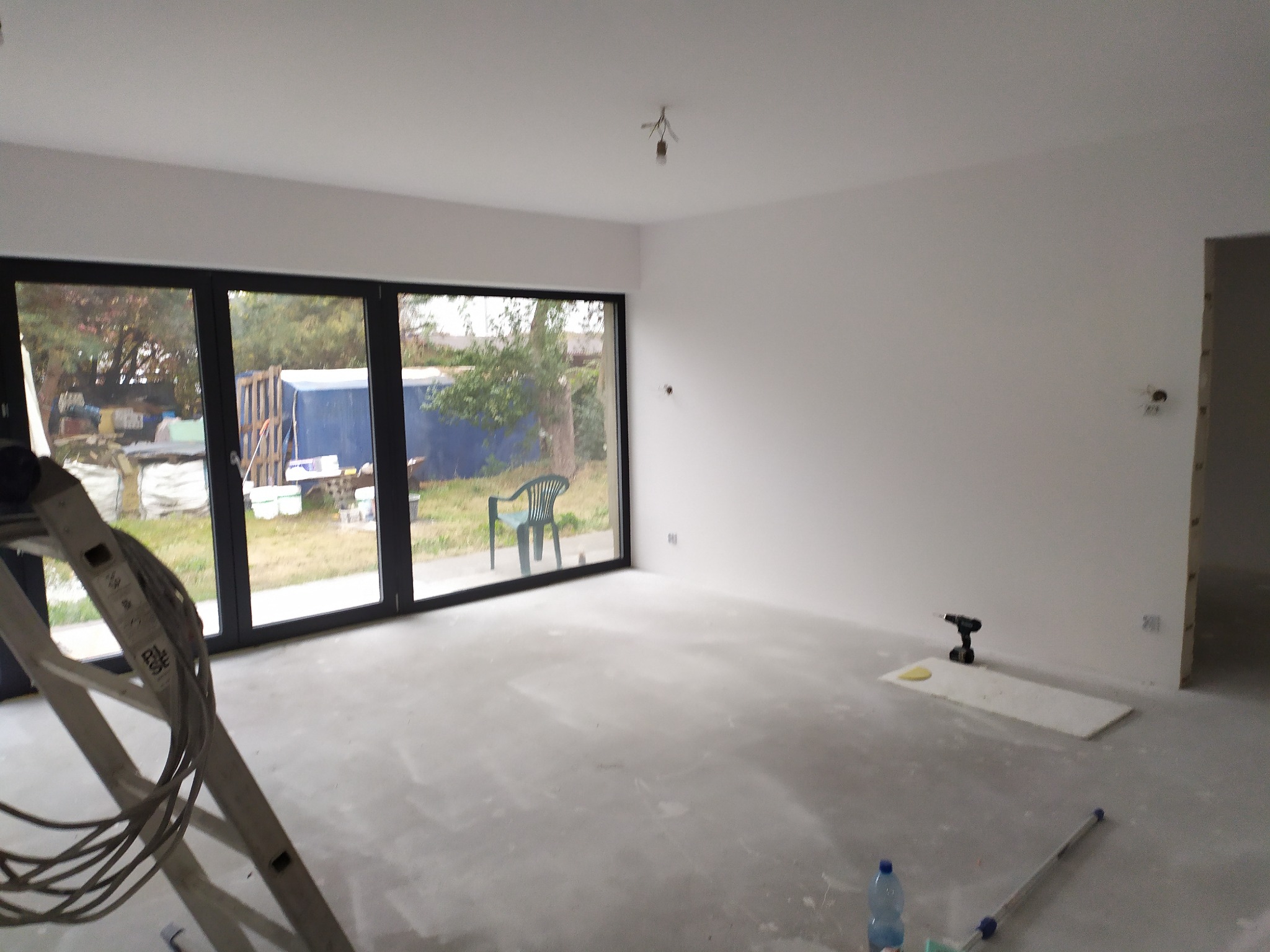 Wnętrze pokoju w trakcie prac wykończeniowych: świeżo pomalowane ściany na biało, surowa podłoga, drabina malarska w rogu, widok na ogród przez duże okno, narzędzia i butelka wody na podłodze.