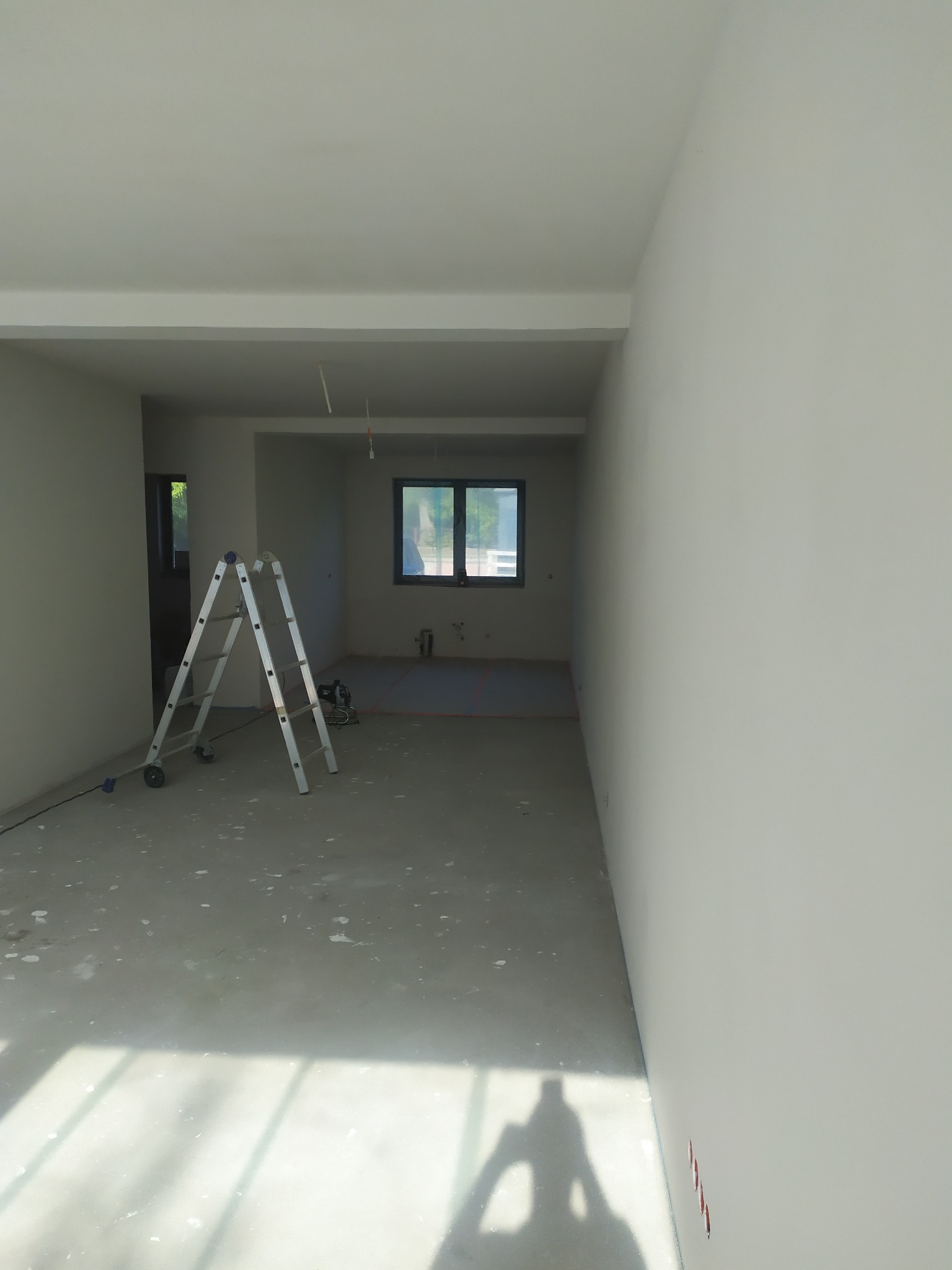 Wnętrze w trakcie prac wykończeniowych: świeżo pomalowane ściany w odcieniach szarości, metalowa drabina rozstawiona w pokoju, widoczne ślady po pracach na podłodze i przygotowane do dalszych działań.