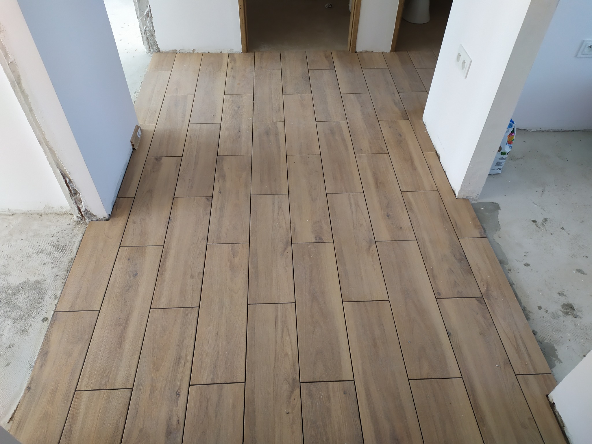 Ułożona podłoga z płytek imitujących drewno, widok z góry, surowe ściany i futryny w tle.