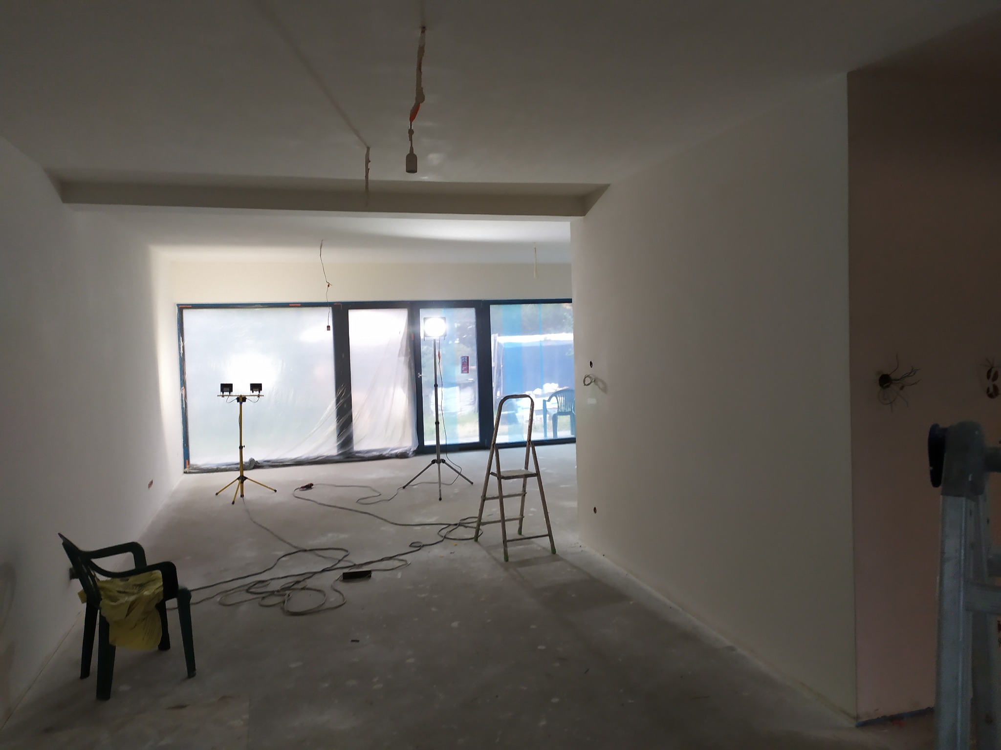 Wnętrze w trakcie prac wykończeniowych: świeżo pomalowane ściany w jasnych odcieniach, zabezpieczone okna folią, drabina, krzesło i lampy budowlane na trójnogach.
