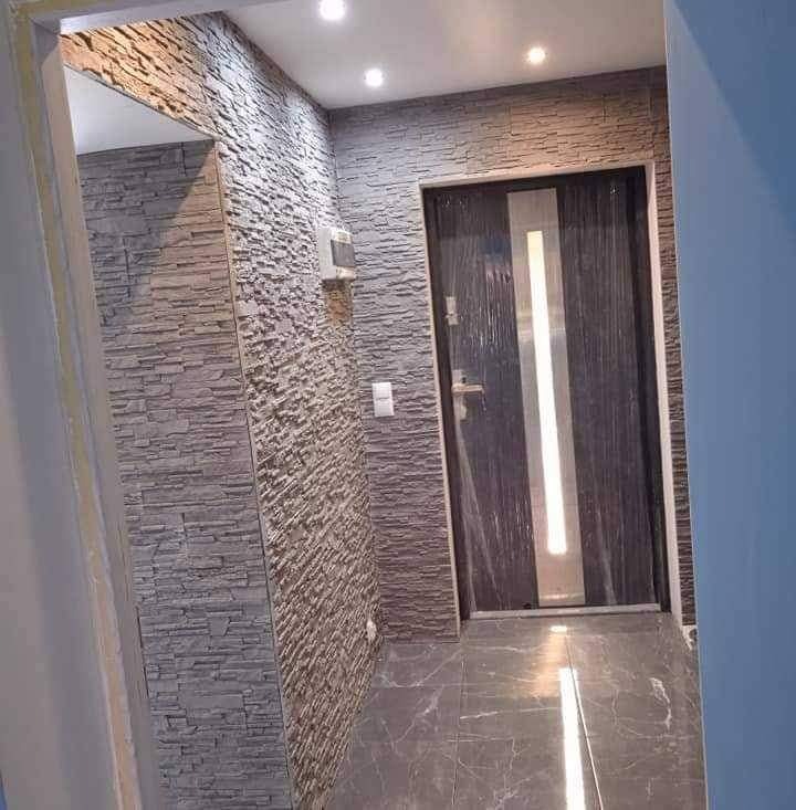 Korytarz z wykończonymi ścianami imitującymi kamień, szara podłoga z marmurkowym wzorem i nowoczesne drzwi wewnętrzne z pionowym, srebrnym paskiem, częściowo osłonięte folią ochronną.