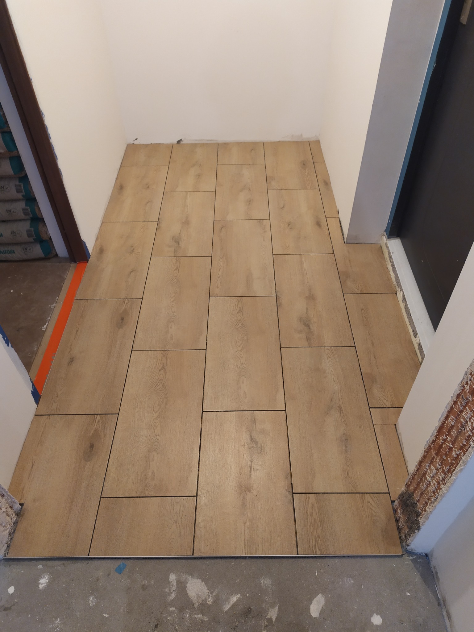 Ułożona podłoga z płytek imitujących drewno w wąskim przedpokoju, widoczne niedokończone obramowania drzwi i ślady po pracach budowlanych.