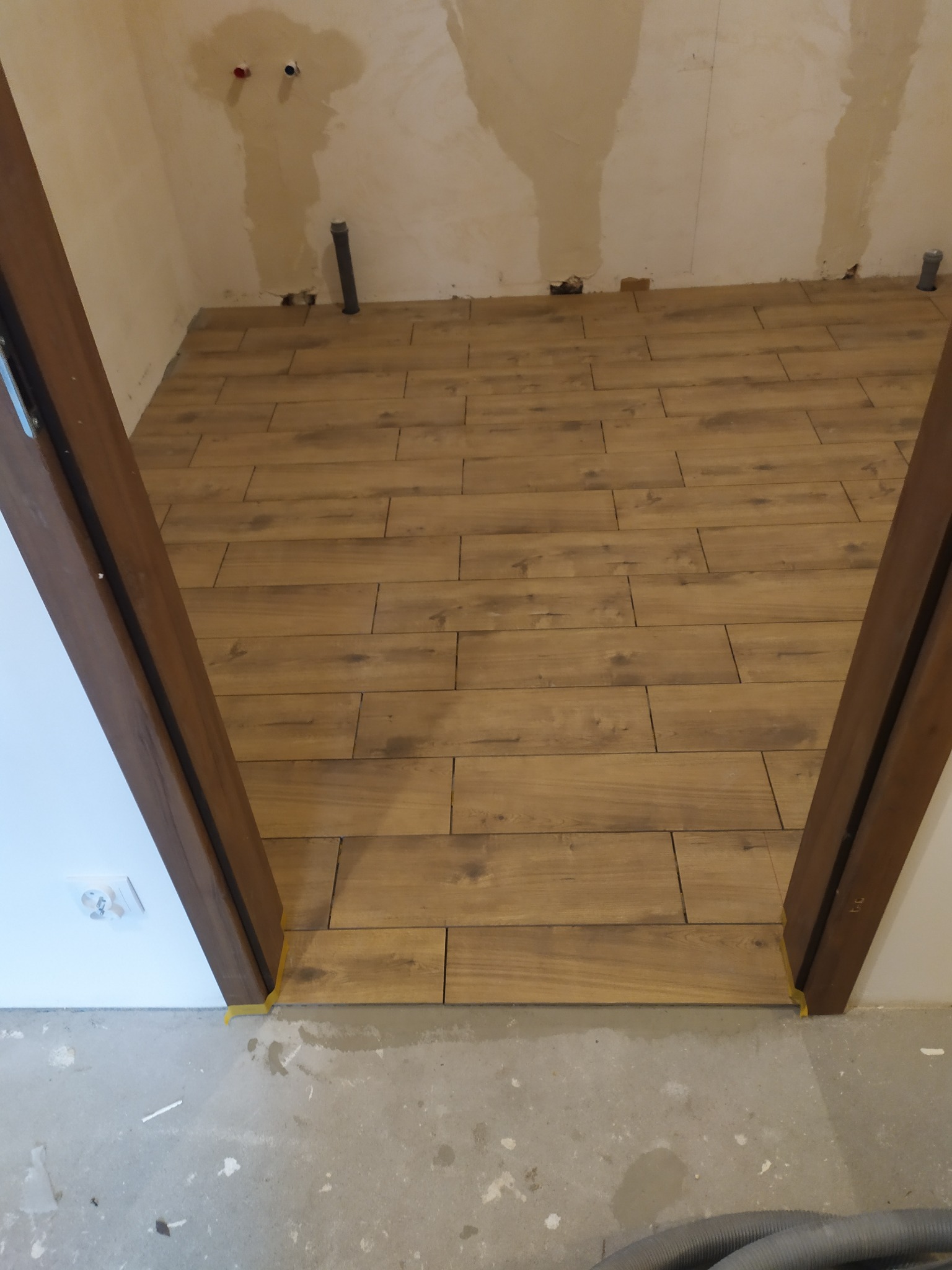 Układanie podłogi z płytek imitujących drewno w pomieszczeniu w trakcie prac wykończeniowych, widoczne niedoskonałości na ścianach i rurki instalacyjne, brązowa ościeżnica drzwiowa z żółtą taśmą...