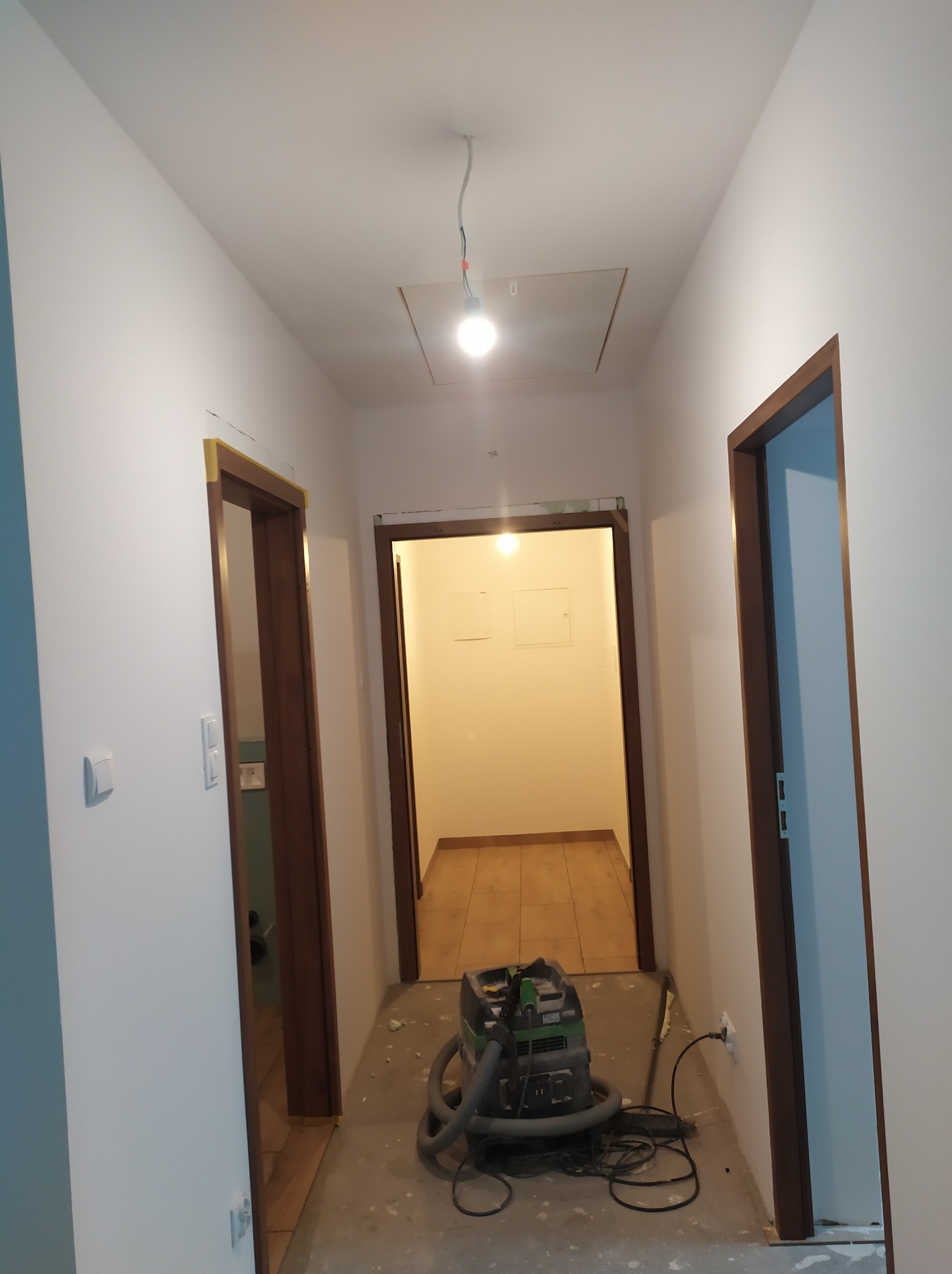 Korytarz w trakcie remontu: świeżo pomalowane białe ściany, brązowe ościeżnice, zabezpieczone żółtą taśmą, odkurzacz przemysłowy na szarym, częściowo zakrytym folią podłożu.