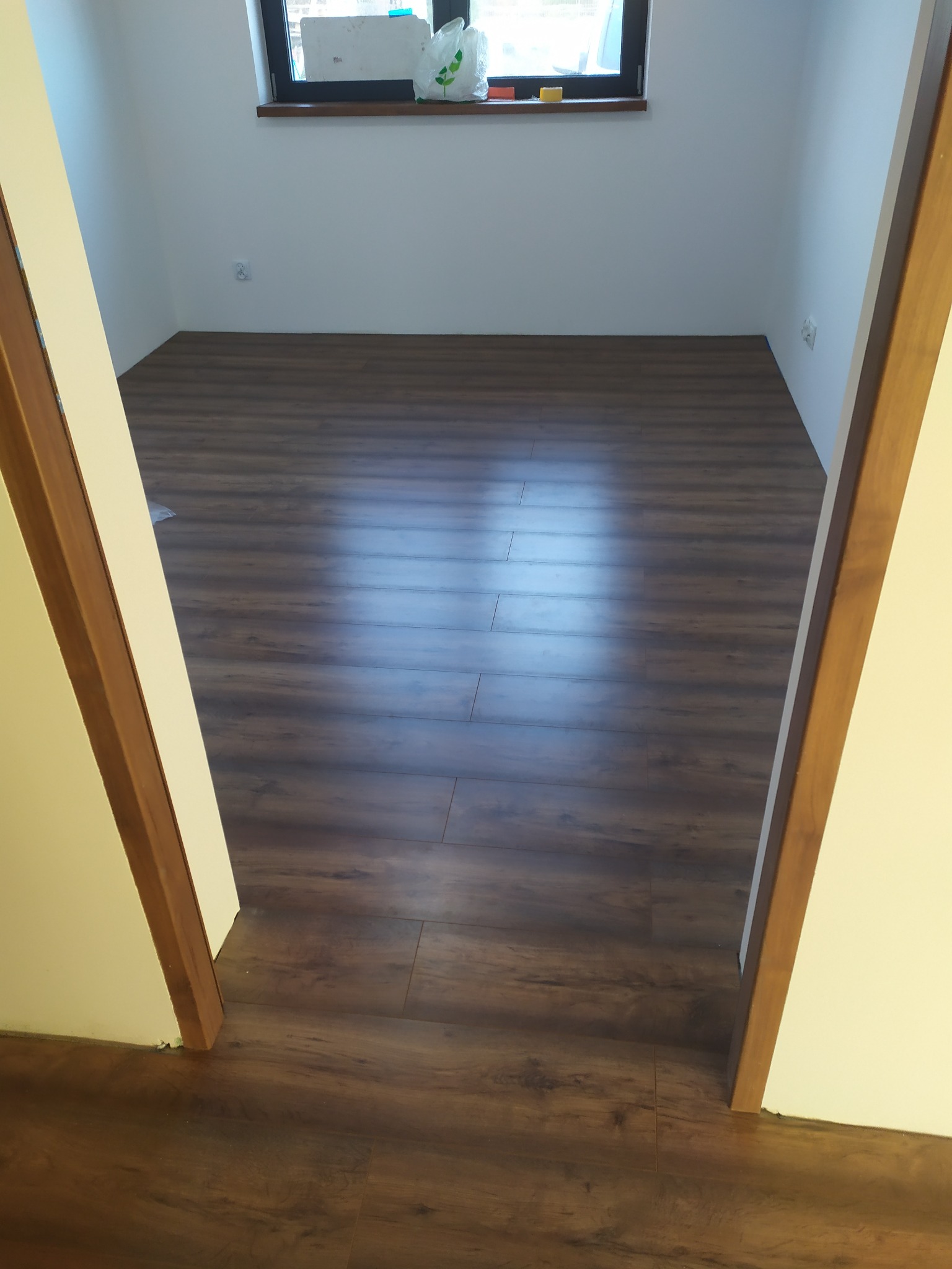 Ułożona podłoga z paneli imitujących drewno w pustym, pomalowanym na biało pokoju, widok zza drewnianej futryny drzwiowej.