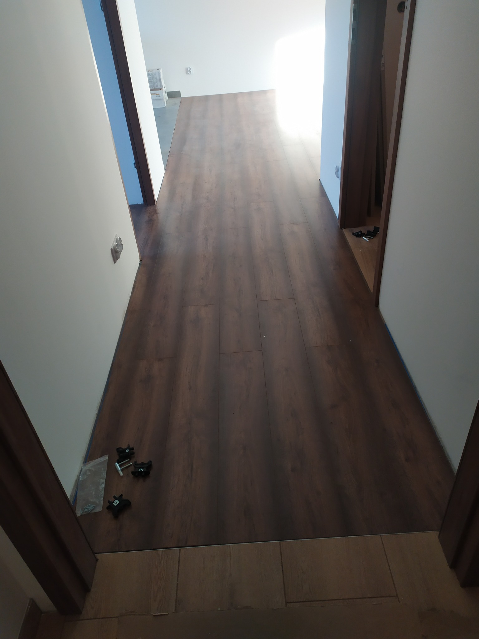 Korytarz z nową, ciemnobrązową podłogą z paneli, widoczne akcesoria montażowe przy drzwiach, świeżo pomalowane ściany na biało.