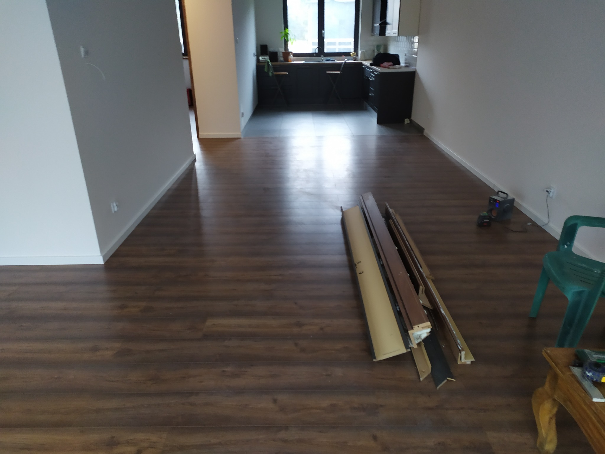 Przygotowanie do montażu paneli podłogowych w pustym pokoju z widoczną kuchnią w tle, narzędzia i listwy przypodłogowe leżą na podłodze.