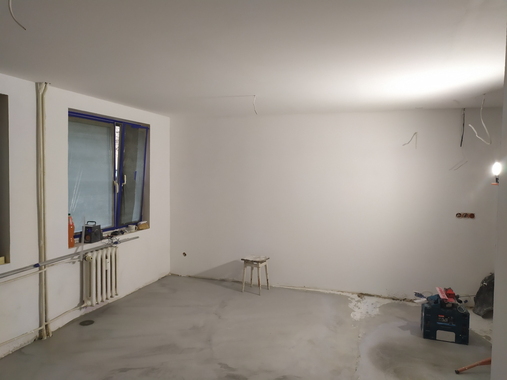 Pokój w trakcie remontu: wylewka na podłodze, świeżo pomalowane ściany, widoczne narzędzia i drabina, okno z niebieską ramą, grzejnik i wystające kable ze ściany.
