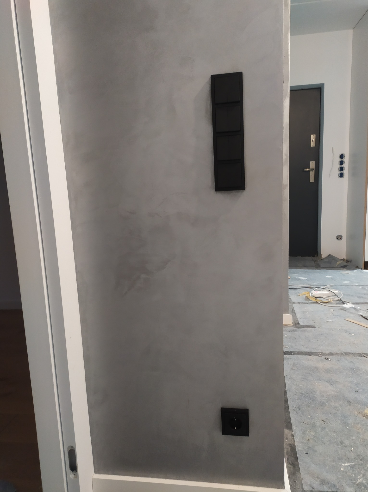 Ściana pokryta szarą, dekoracyjną gładzią w mieszkaniu, z zamontowanym czarnym włącznikiem światła i gniazdkiem elektrycznym; widoczny fragment białej ościeżnicy drzwiowej i czarne drzwi wejściowe...