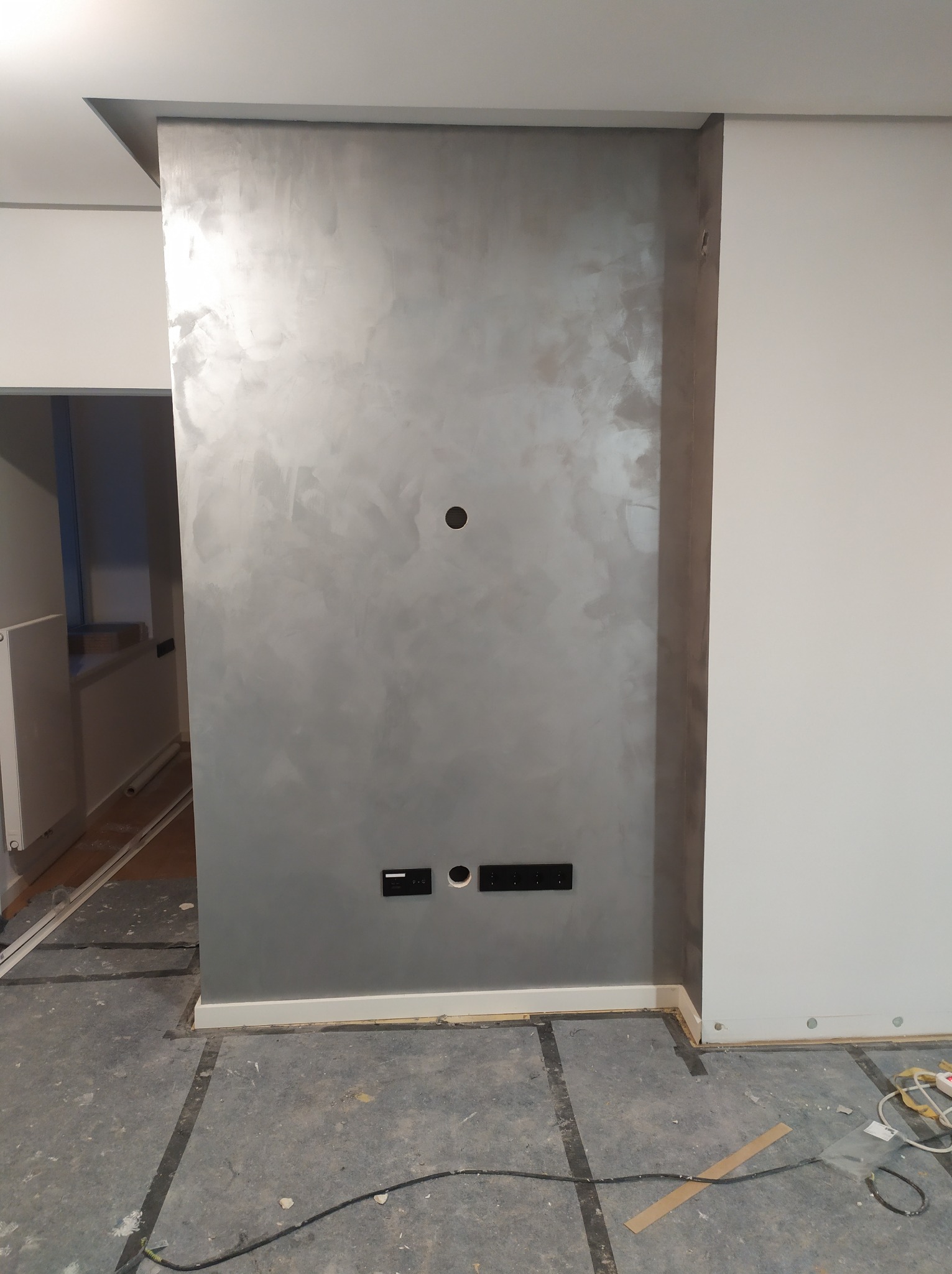 Ściana z dekoracyjną, srebrzystą fakturą w trakcie prac wykończeniowych, widoczne gniazdka elektryczne, otwór na kable oraz zabezpieczona podłoga.