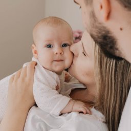 Natalia Cisłowska Fotografia - Mama całuje policzek niemowlęcia o błękitnych oczach, trzymanego na rękach, w tle widoczny fragment brody i szyi taty.