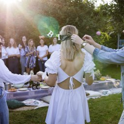 Natalia Cisłowska Fotografia - Uczestniczki wieczoru panieńskiego w ogrodzie: jedna zawiązuje opaskę na oczy przyszłej pannie młodej, w tle grupa kobiet przy stole piknikowym.