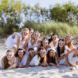 Natalia Cisłowska Fotografia - Grupa kobiet w białych strojach i okularach w kształcie serc z napisem 'Wieczór Panieński' pozuje na piaszczystej plaży, w tle trawy i drzewa.