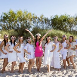 Natalia Cisłowska Fotografia - Zdjęcie grupy kobiet w białych sukienkach i okularach w kształcie serc z napisem 'Bride to be' na plaży, jedna kobieta w różowej sukience podnosi ręce w geście radości, w tle wydmy i drzewa.