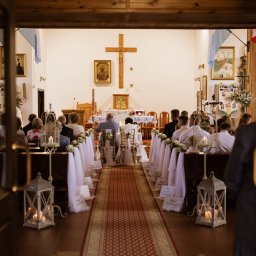 Natalia Cisłowska Fotografia - Widok z zewnątrz na wnętrze kościoła podczas ceremonii ślubnej, z dekoracjami kwiatowymi i lampionami wzdłuż przejścia.