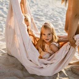 Natalia Cisłowska Fotografia - Dwie małe dziewczynki w pastelowym, dzierganym hamaku na plaży, trzymanym przez dorosłych; jedna dziewczynka patrzy w obiektyw, druga częściowo schowana.