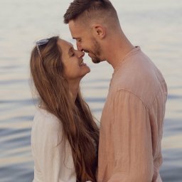 Natalia Cisłowska Fotografia - Zakochana para, mężczyzna z brodą w różowej koszuli i kobieta z okularami na głowie w białej sukience, przytuleni nosem do nosa na tle morza o zachodzie słońca.