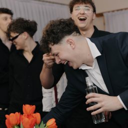 Natalia Cisłowska Fotografia - Rozbawieni młodzi mężczyźni w eleganckich strojach, jeden trzyma butelkę, w tle kwiaty, jasne wnętrze. Uśmiechy i radosna atmosfera.