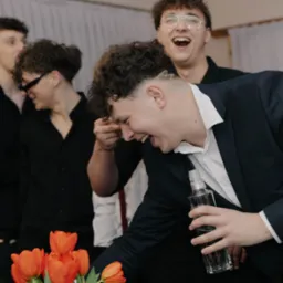 Rozbawieni młodzi mężczyźni w eleganckich strojach, jeden trzyma butelkę, w tle kwiaty, jasne wnętrze. Uśmiechy i radosna atmosfera.