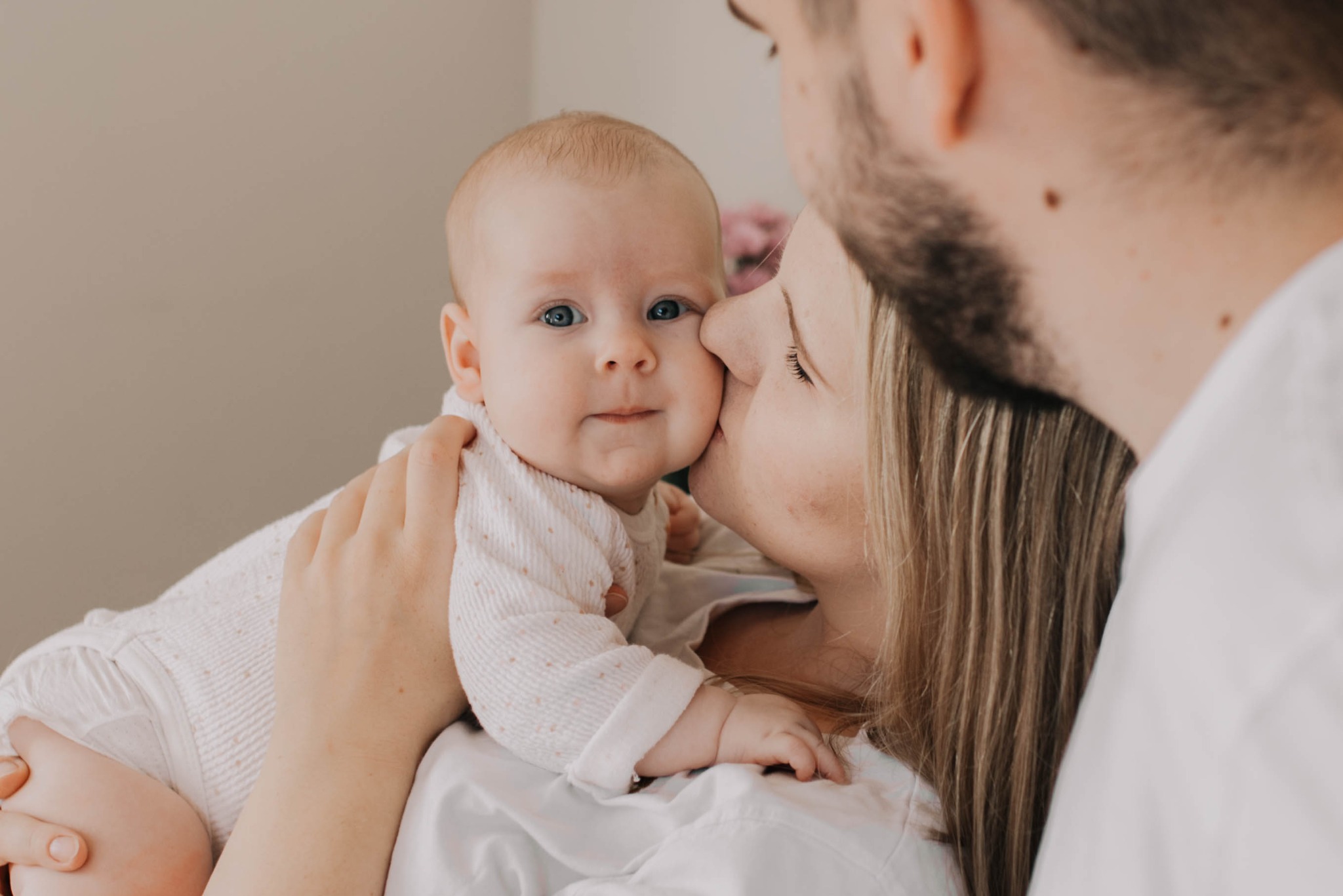 Mama całuje policzek niemowlęcia o błękitnych oczach, trzymanego na rękach, w tle widoczny fragment brody i szyi taty.
