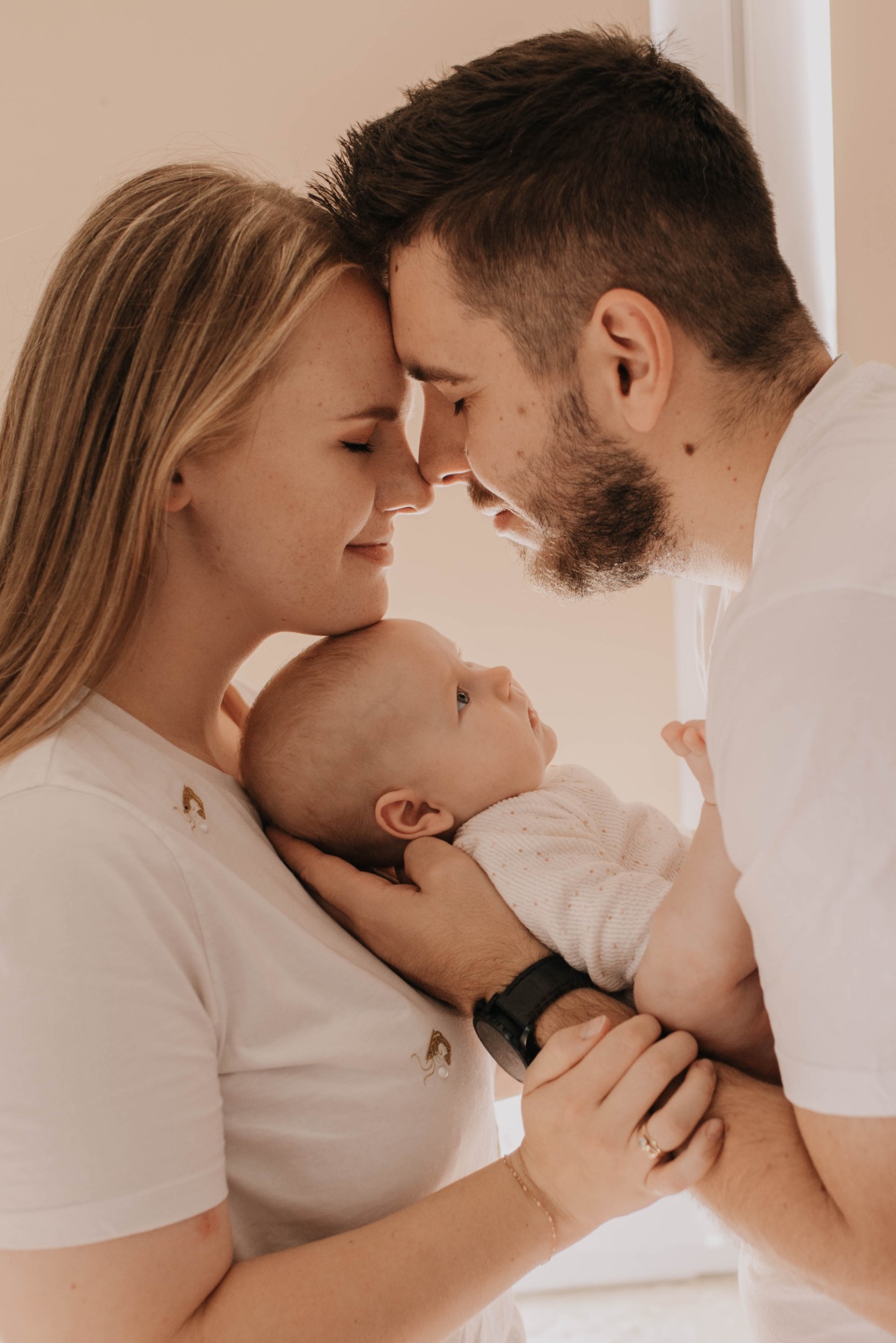 Rodzinne ujęcie w Orlu: mama, tata i niemowlę tulą się, dotykając czołami w ciepłym, jasnym świetle. Na białych koszulkach rodziców delikatny haft.