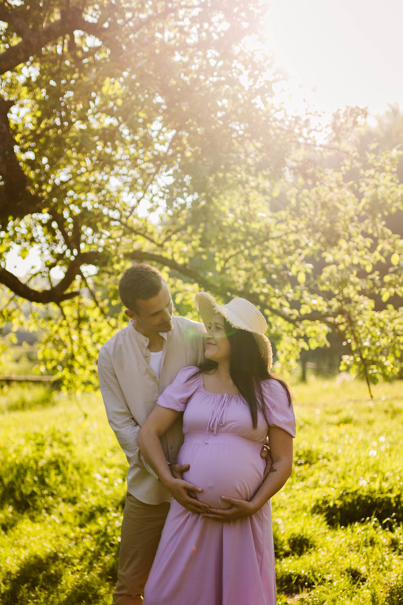Przyszła mama w liliowej sukience i słomkowym kapeluszu, czule obejmowana przez partnera na tle zielonego ogrodu w promieniach słońca.