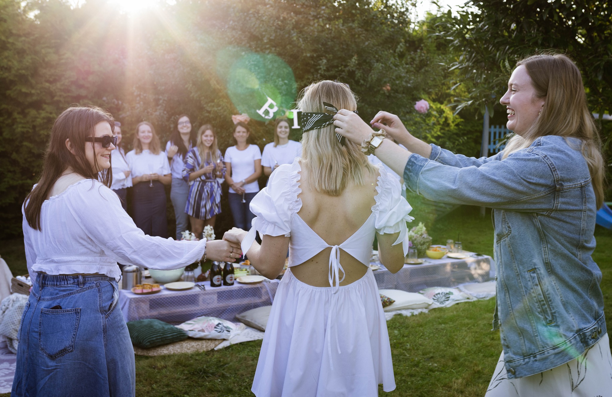 Uczestniczki wieczoru panieńskiego w ogrodzie: jedna zawiązuje opaskę na oczy przyszłej pannie młodej, w tle grupa kobiet przy stole piknikowym.