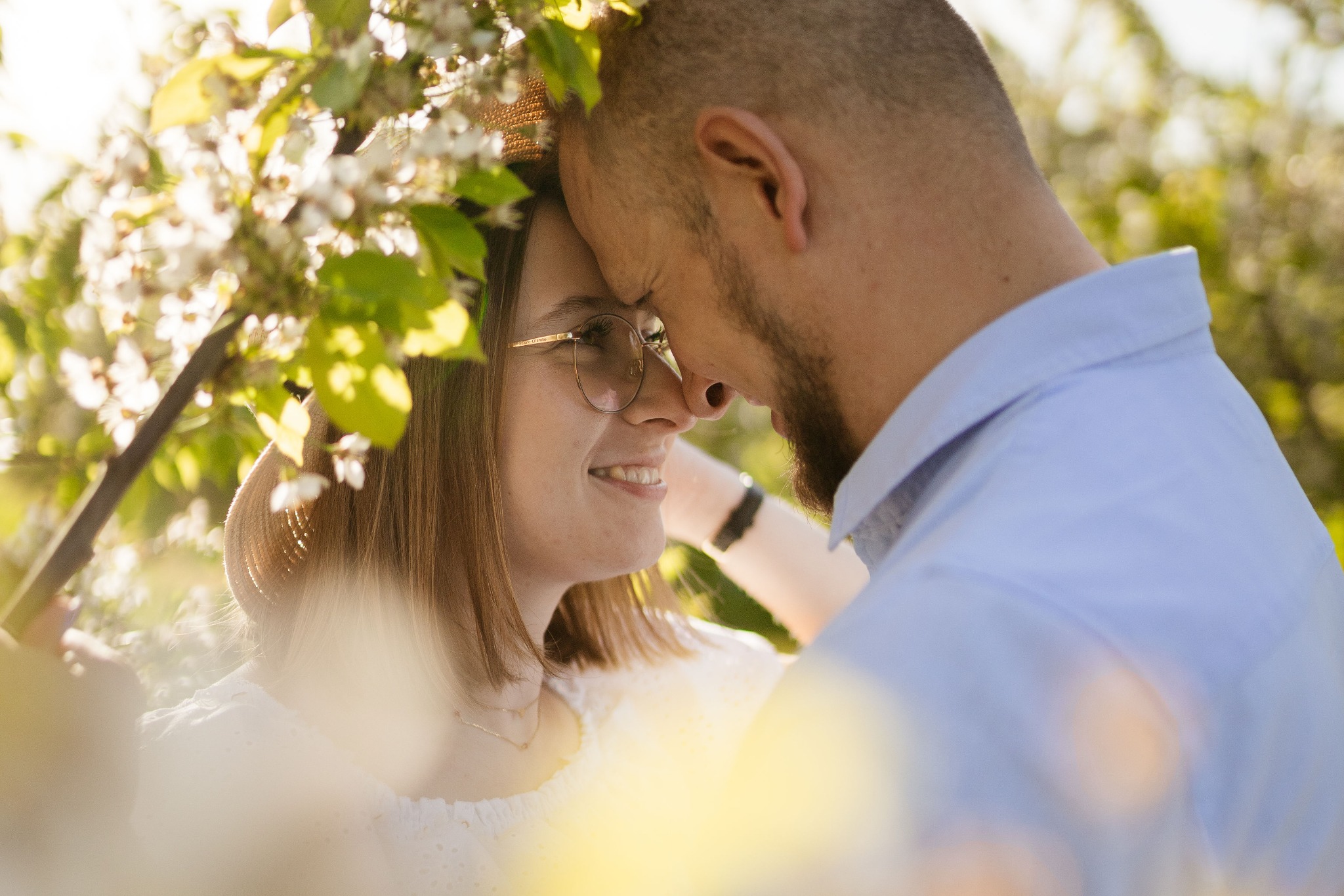 Zakochana para w wiosennym sadzie, twarze blisko siebie, ona w okularach i kapeluszu, on z brodą, otoczeni kwitnącymi gałęziami.