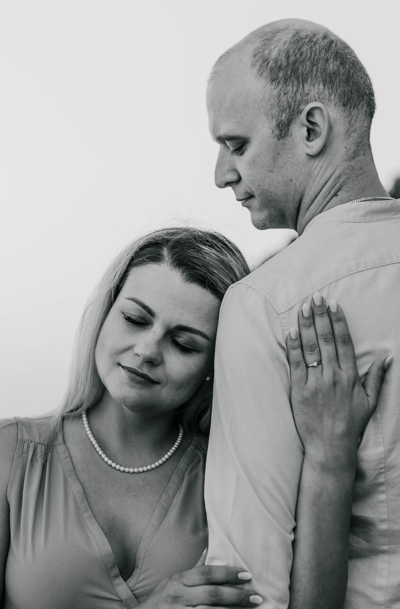 Czarno-białe zbliżenie pary: kobieta opiera głowę o ramię mężczyzny, delikatny uśmiech, perłowy naszyjnik, pierścionek zaręczynowy na dłoni spoczywającej na jego ramieniu.