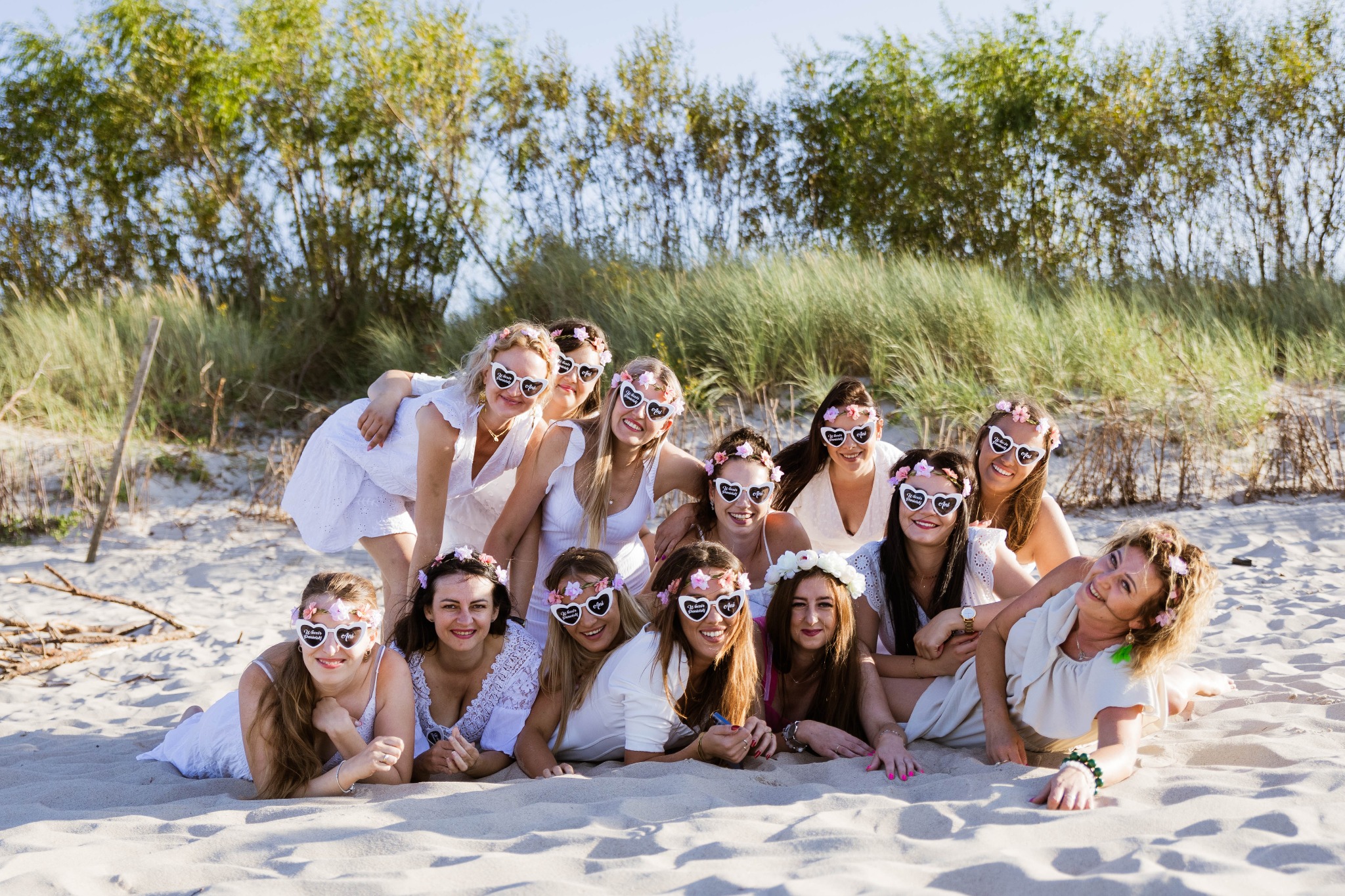 Grupa kobiet w białych strojach i okularach w kształcie serc z napisem 'Wieczór Panieński' pozuje na piaszczystej plaży, w tle trawy i drzewa.