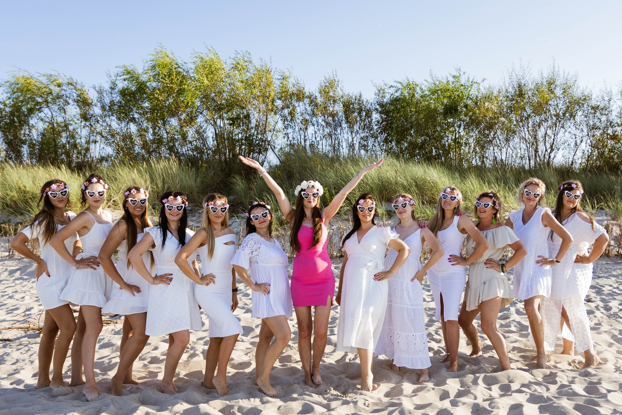 Zdjęcie grupy kobiet w białych sukienkach i okularach w kształcie serc z napisem 'Bride to be' na plaży, jedna kobieta w różowej sukience podnosi ręce w geście radości, w tle wydmy i drzewa.