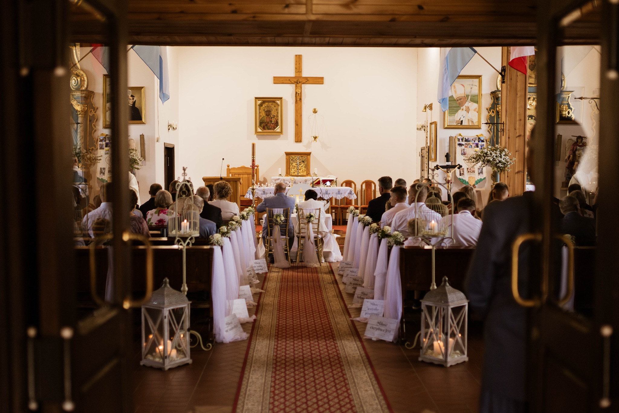 Widok z zewnątrz na wnętrze kościoła podczas ceremonii ślubnej, z dekoracjami kwiatowymi i lampionami wzdłuż przejścia.