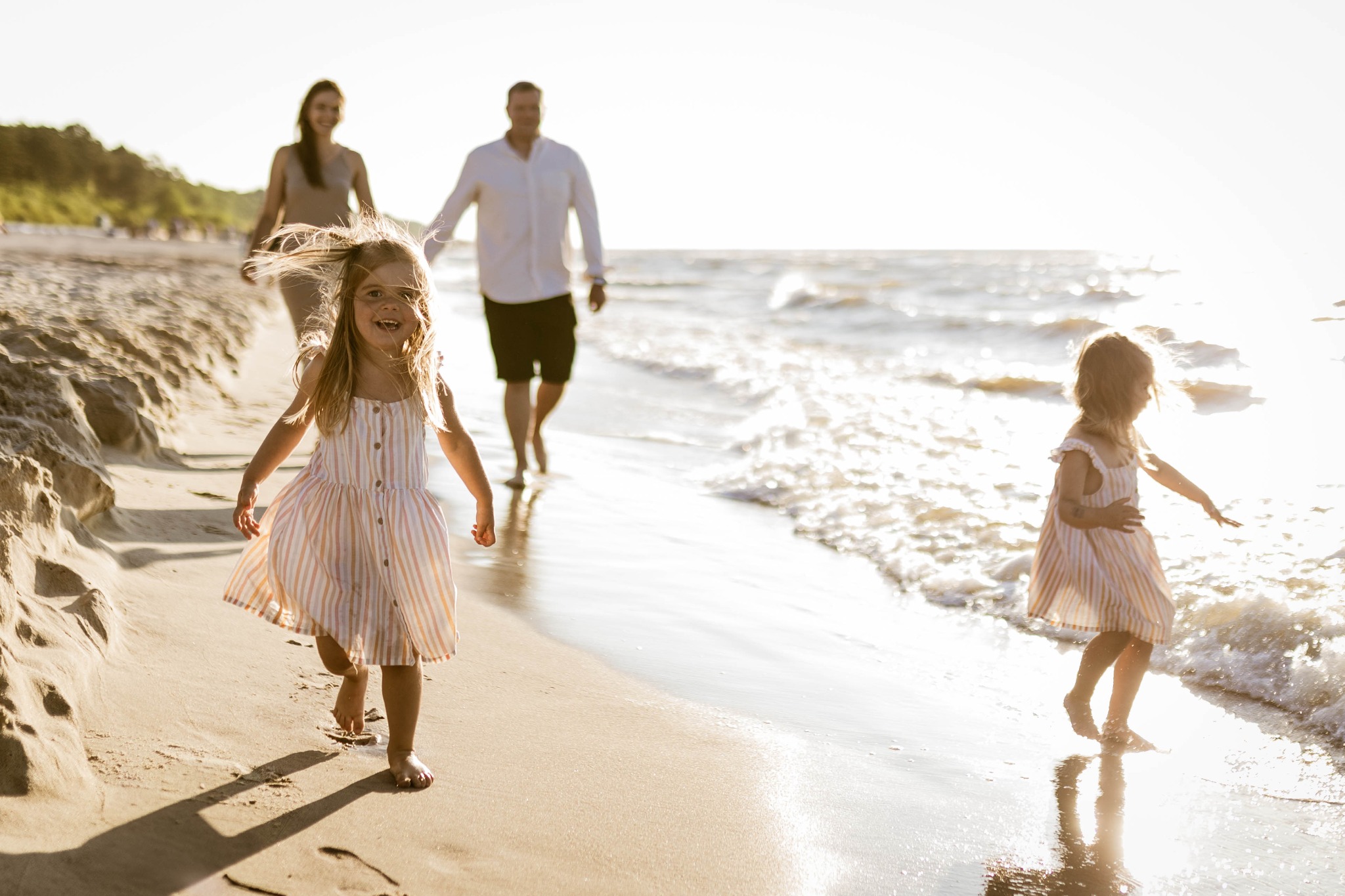 Dziewczynka w paski biegnie po plaży w kierunku aparatu, rodzice i druga dziewczynka w tle, promienie słońca odbijają się w wodzie.