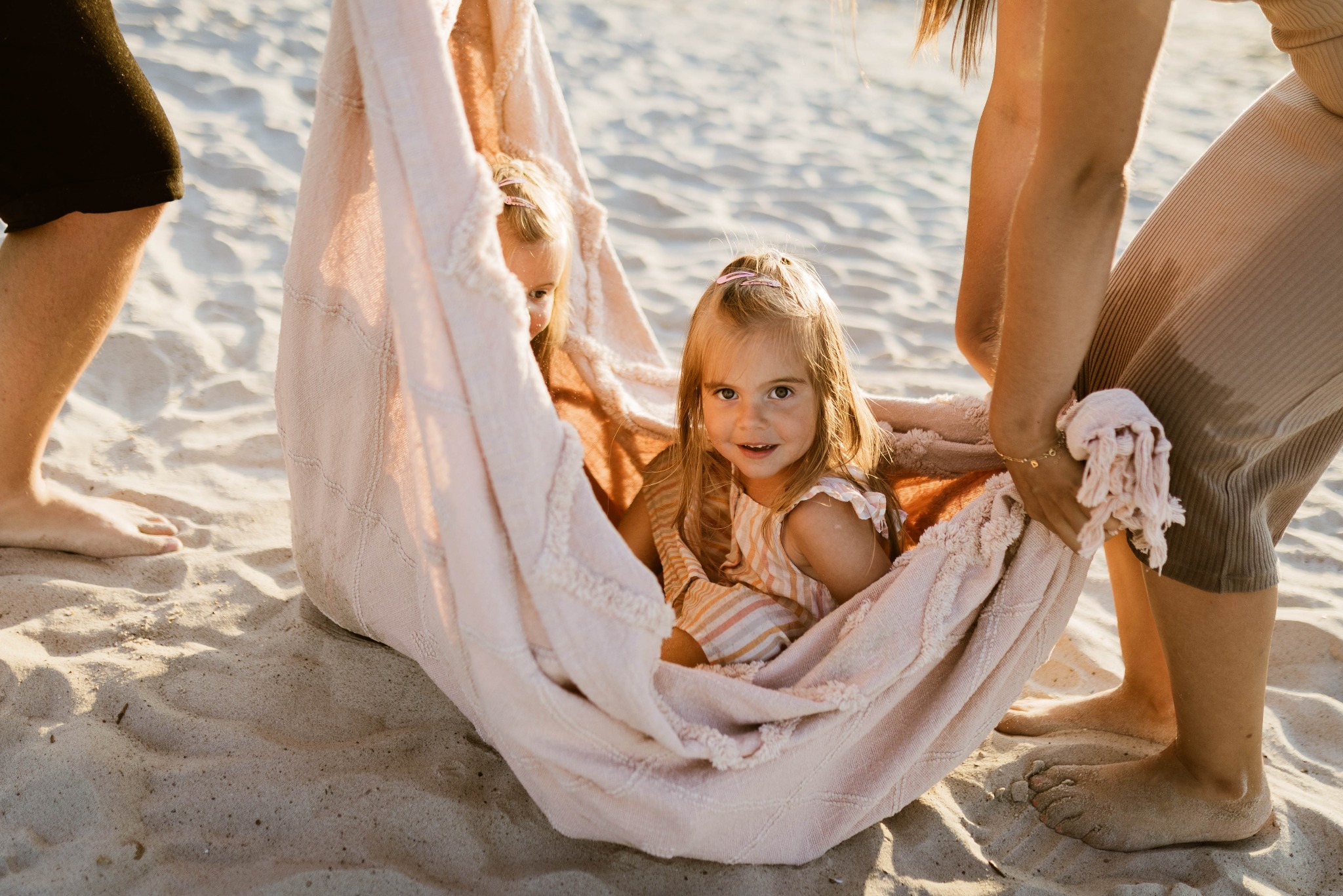 Dwie małe dziewczynki w pastelowym, dzierganym hamaku na plaży, trzymanym przez dorosłych; jedna dziewczynka patrzy w obiektyw, druga częściowo schowana.