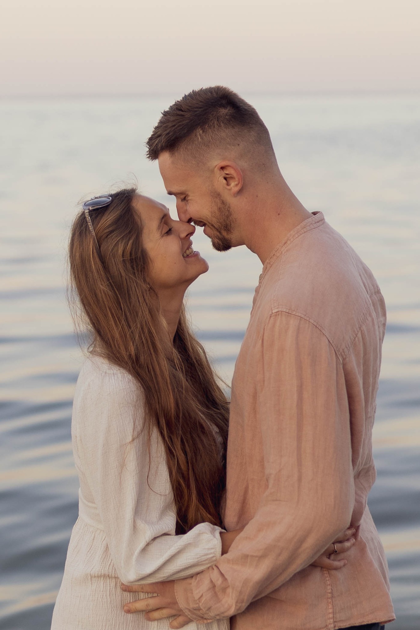 Zakochana para, mężczyzna z brodą w różowej koszuli i kobieta z okularami na głowie w białej sukience, przytuleni nosem do nosa na tle morza o zachodzie słońca.