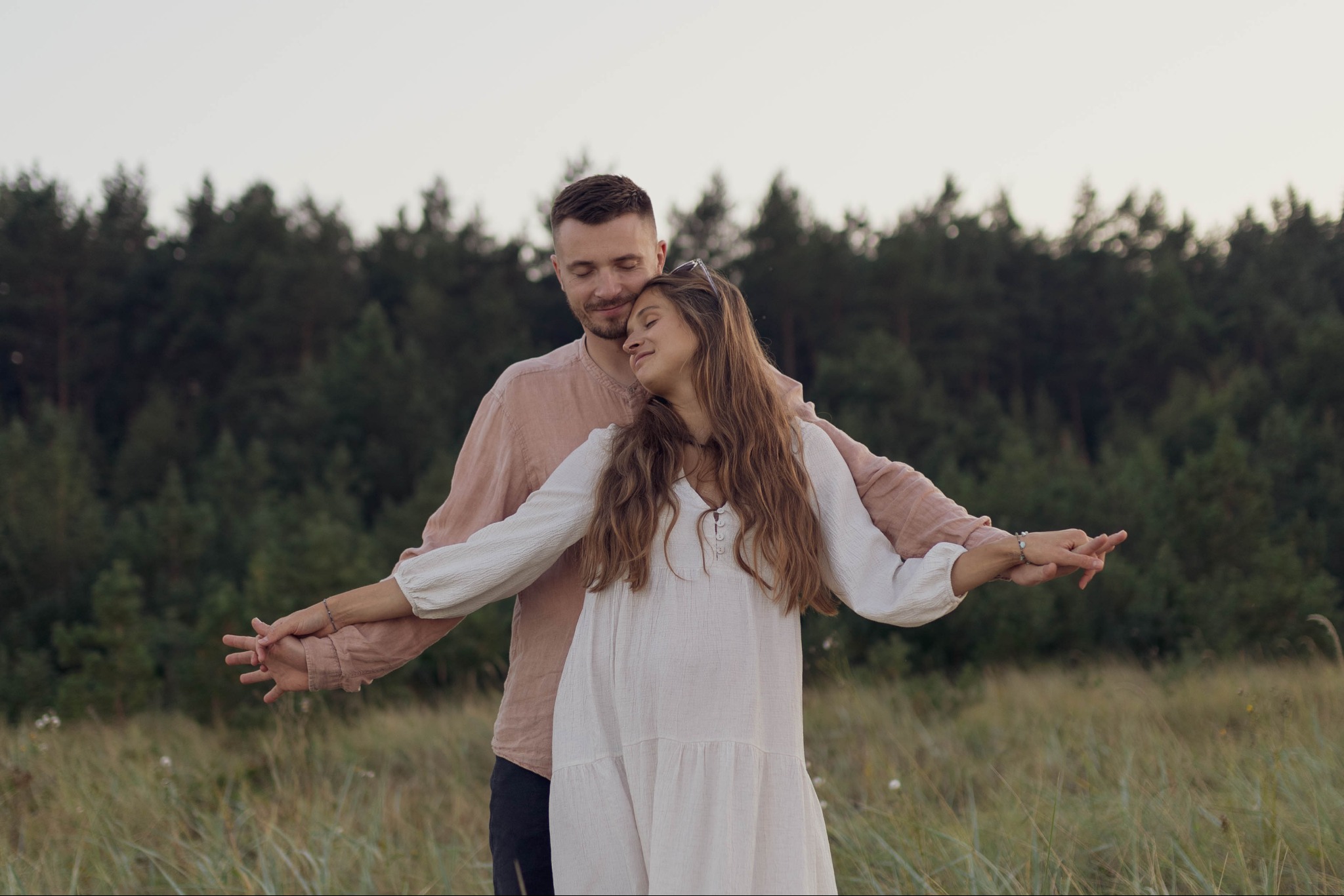 Młoda para w objęciach na tle lasu i łąki, kobieta w zwiewnej sukience z rozłożonymi ramionami, mężczyzna w koszuli z lnu, oboje z zamkniętymi oczami w delikatnym uśmiechu.