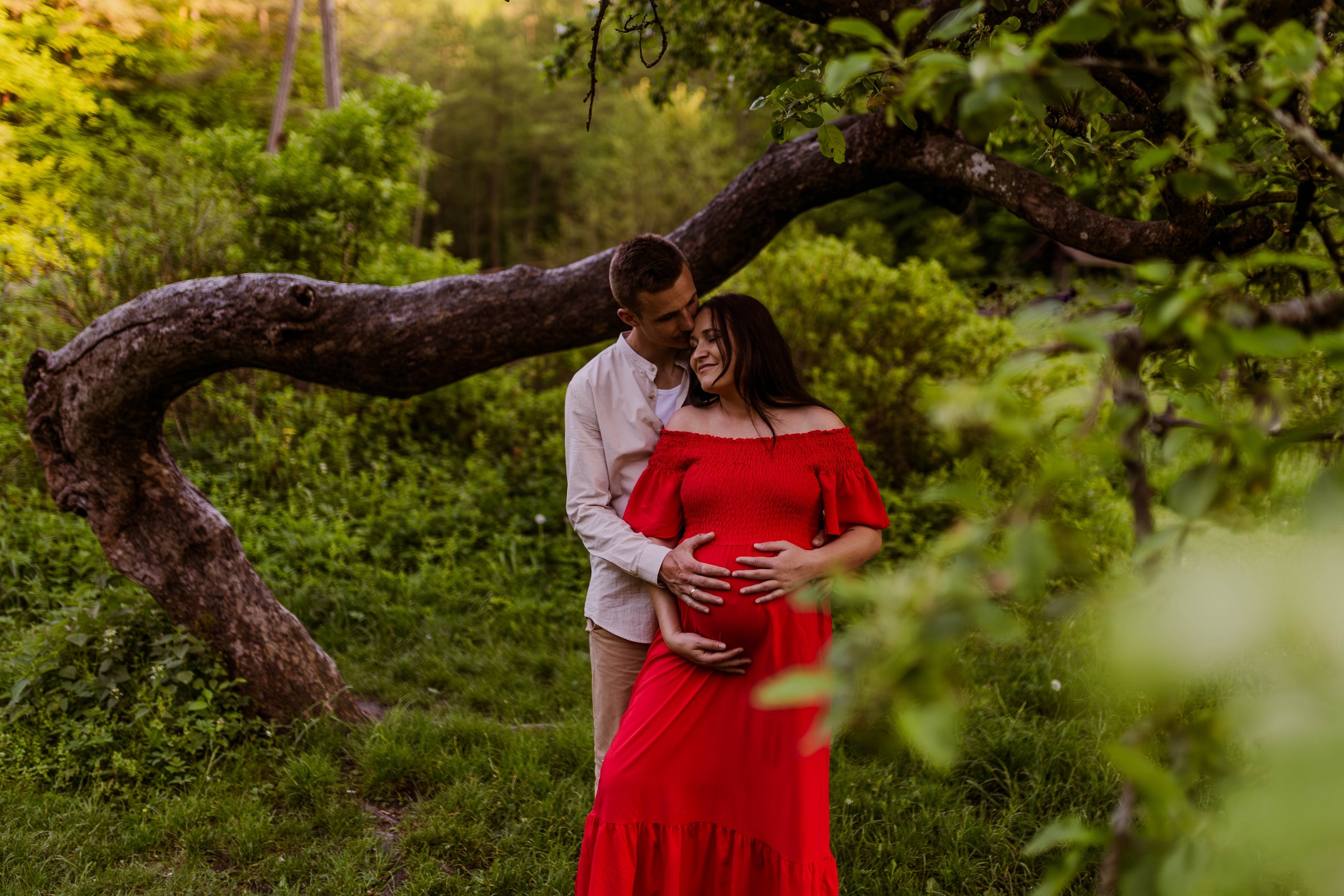 Przyszła mama w czerwonej sukni, obejmowana przez partnera na tle zieleni i malowniczego, rozłożystego drzewa.