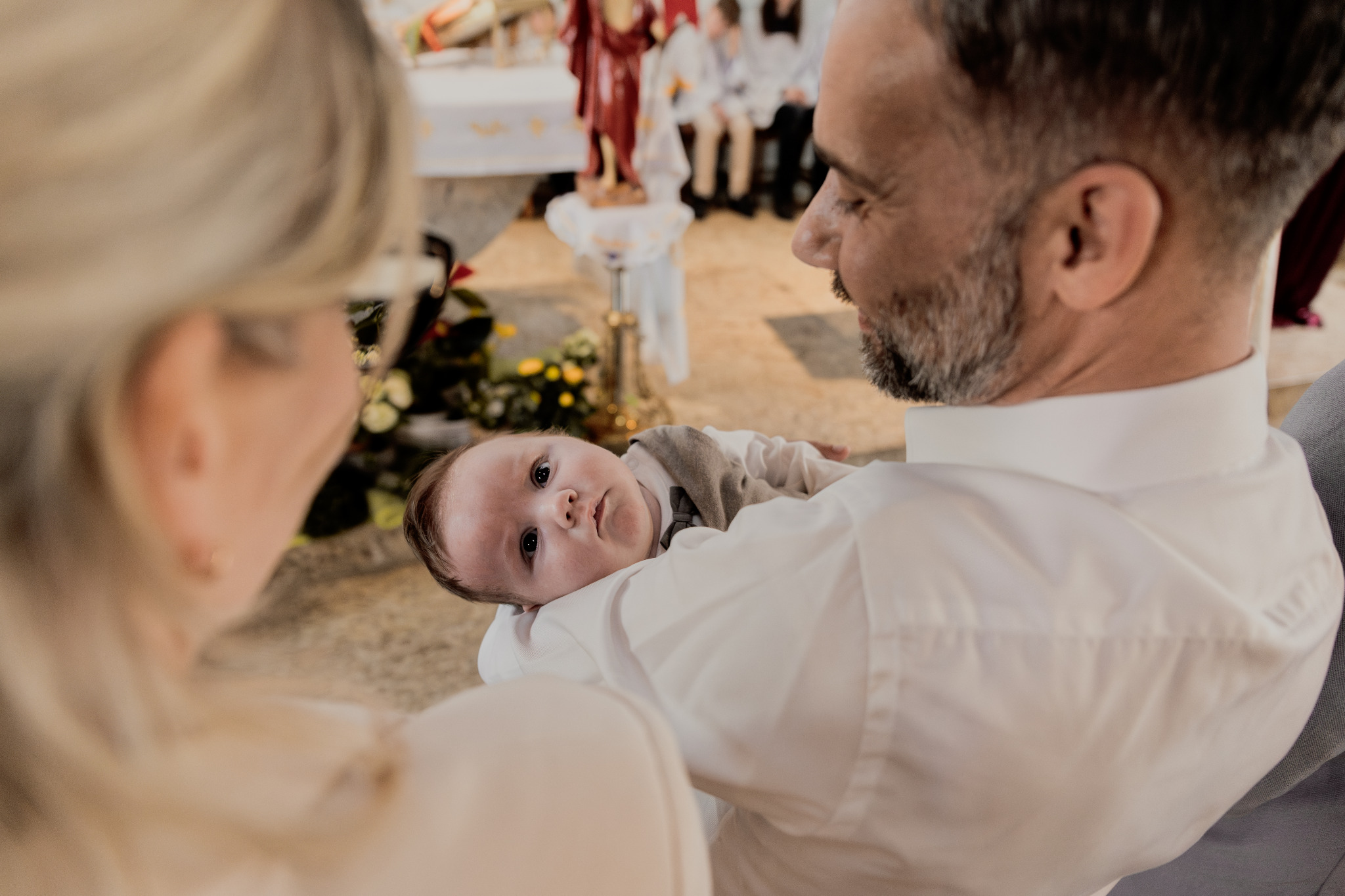 Uśmiechnięty ojciec trzyma na rękach niemowlę w kościele, spoglądające w obiektyw, z widoczną matką z boku i ołtarzem w tle. Jasne, naturalne światło.