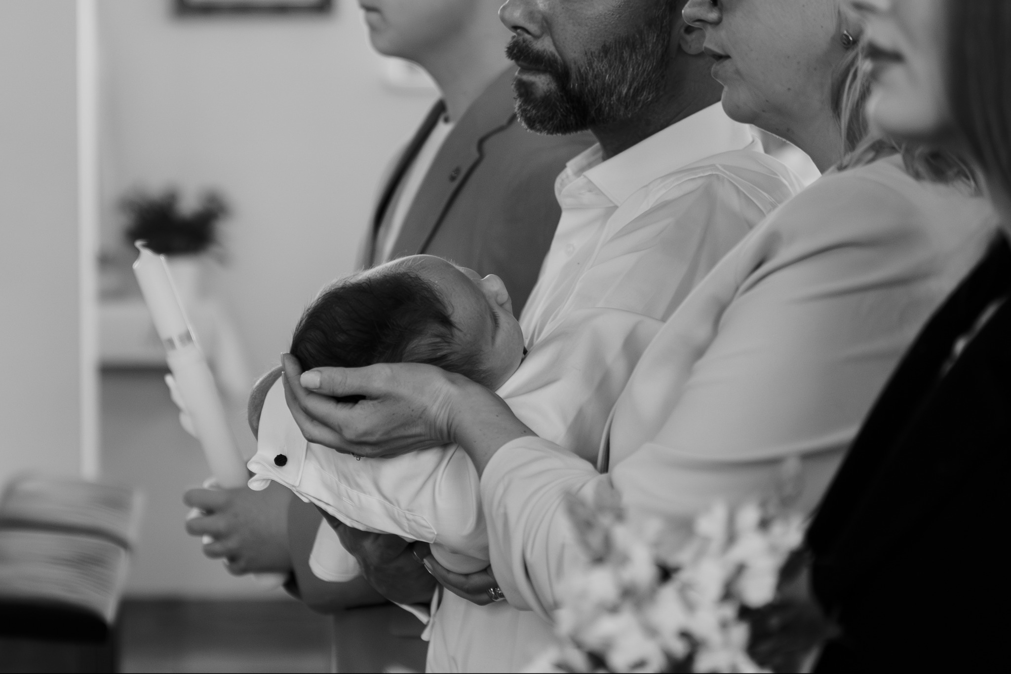 Czarno-białe zdjęcie z uroczystości chrztu: mężczyzna trzyma śpiące niemowlę, obok osoby z zapalonymi świecami i bukietem kwiatów. Delikatne światło.