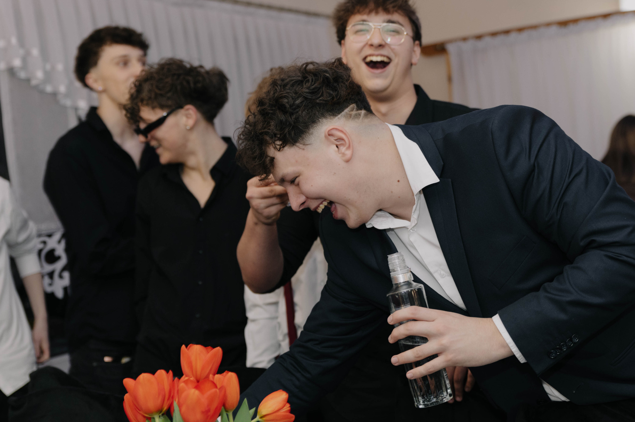 Rozbawieni młodzi mężczyźni w eleganckich strojach, jeden trzyma butelkę, w tle kwiaty, jasne wnętrze. Uśmiechy i radosna atmosfera.