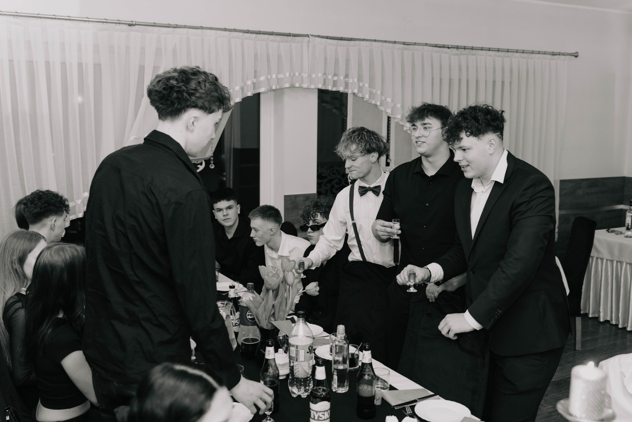 Czarno-białe zdjęcie grupy młodych ludzi w eleganckich strojach, w trakcie uroczystości w Orlu. Widoczne stoły z napojami i kwiatami, tło zasłonięte białymi firankami.