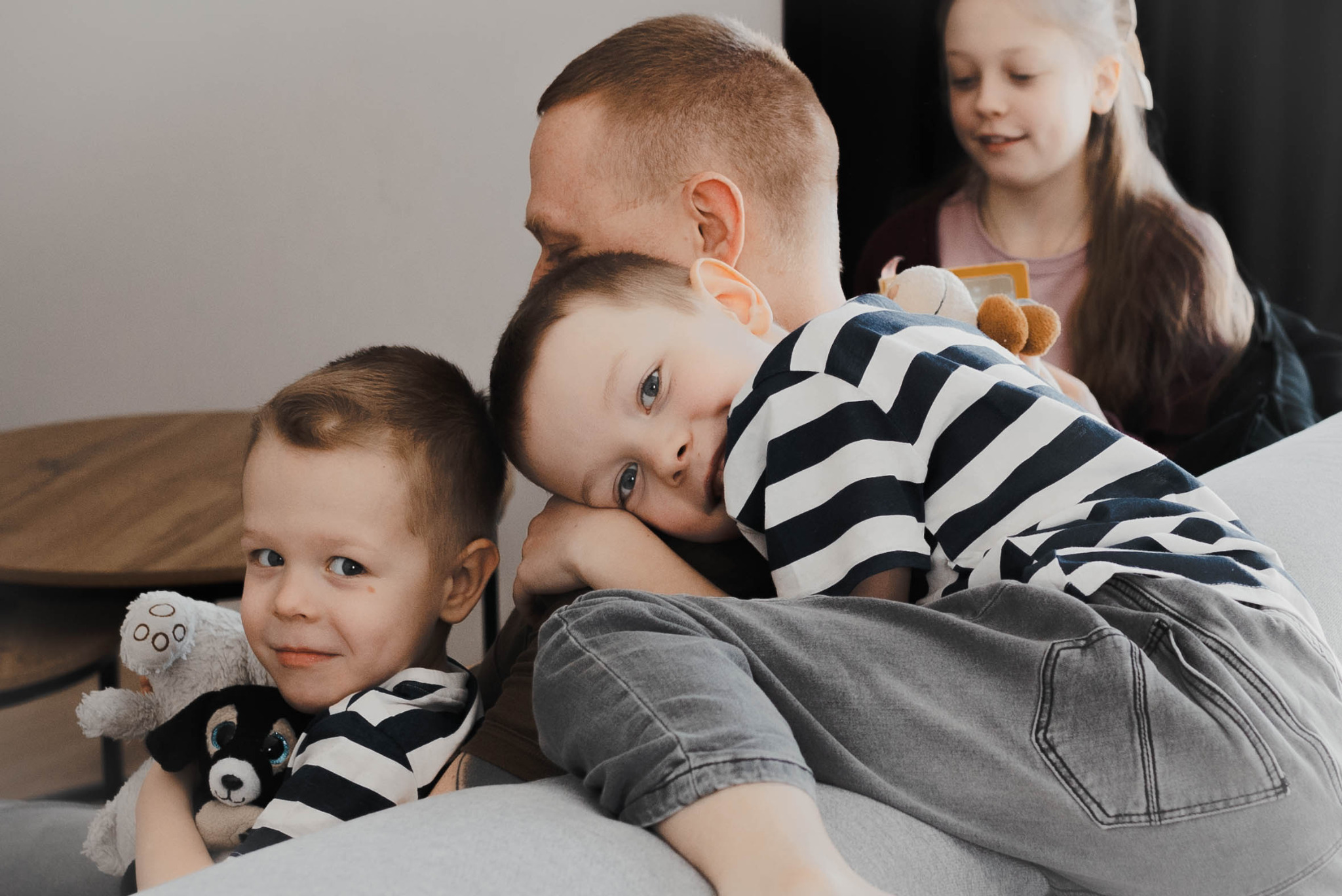 Tata z trójką dzieci na kanapie. Chłopiec w paski opiera się o tatę, drugi trzyma pluszaki, a dziewczynka czyta książkę w tle. Ciepłe, naturalne światło.