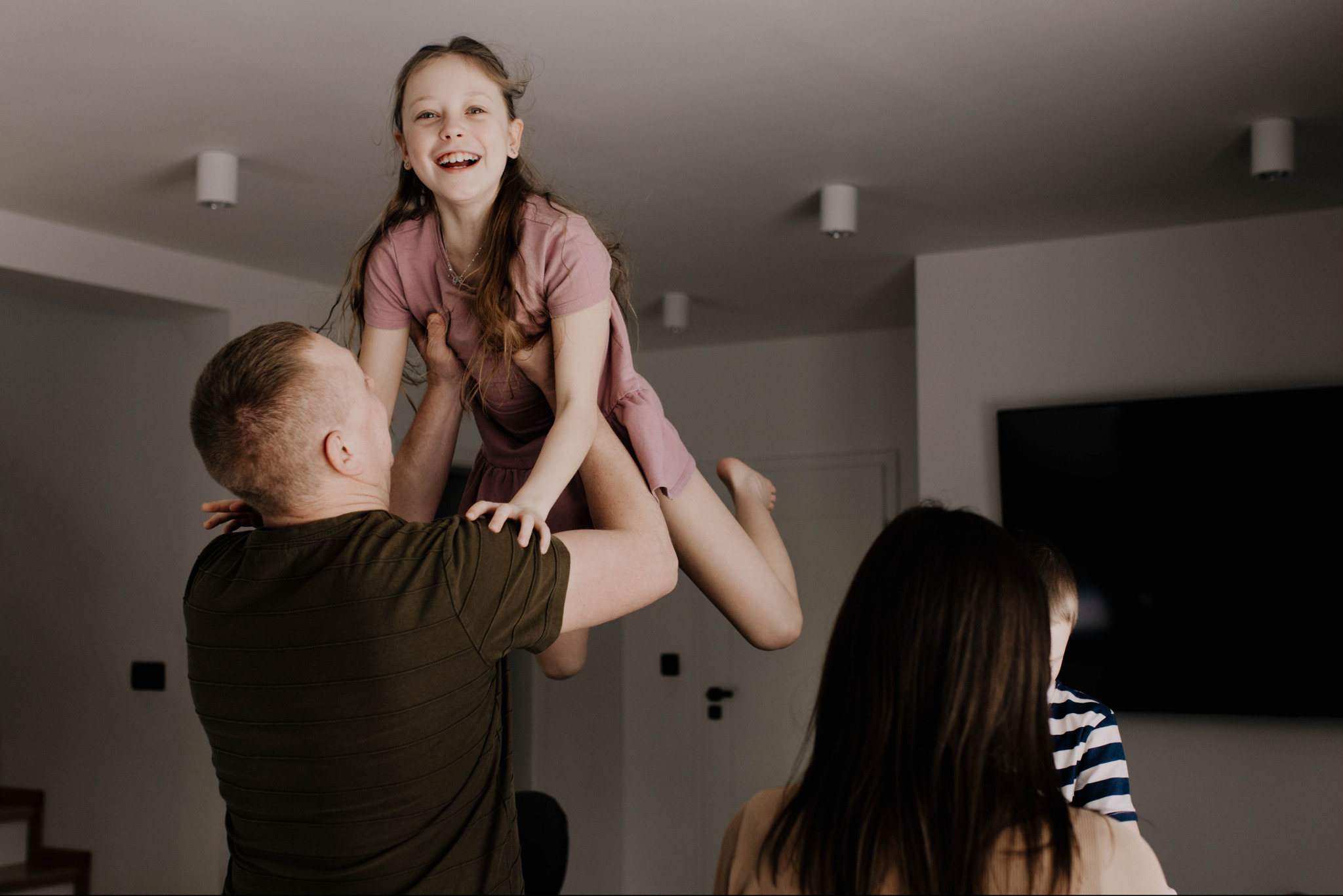 Tata podnosi uśmiechniętą córkę w różowej sukience, w tle mama i syn w salonie z telewizorem. Ciepłe, naturalne światło.