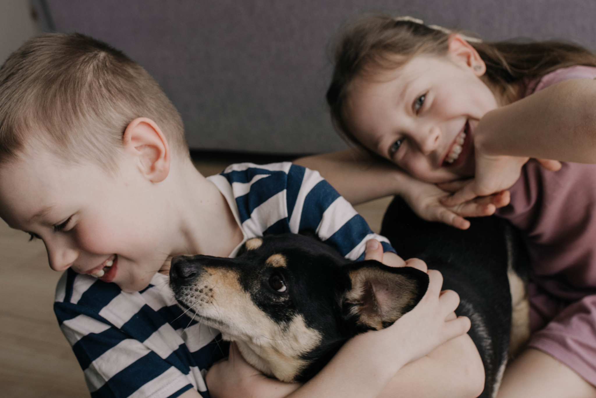 Uśmiechnięte rodzeństwo obejmuje psa rasy mieszanej w domowym wnętrzu. Chłopiec w bluzie w paski i dziewczynka w różowej koszulce. Ciepłe, naturalne światło.